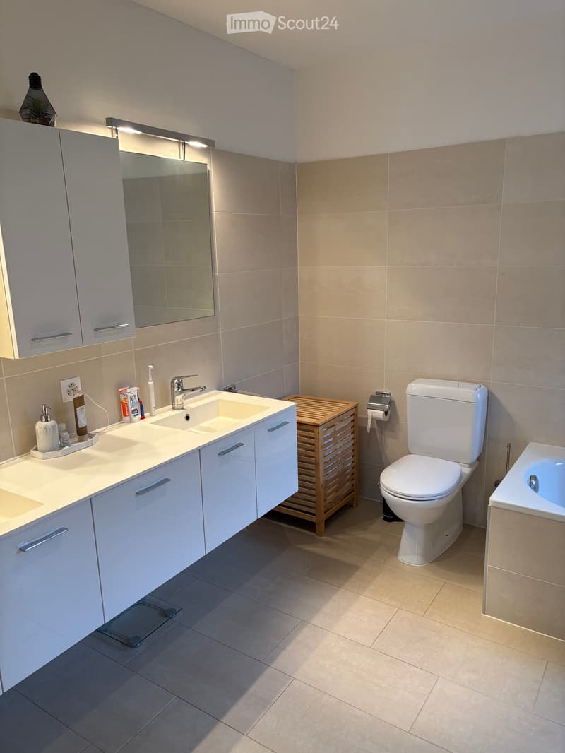 Modern bathroom with white cabinets, double sink, toilet, bathtub, and a wooden shelf. Features a large mirror with built-in lighting and various toiletries on the sink counter.