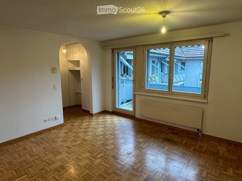 Empty room with wooden parquet flooring, white walls, large window with view to balcony, radiator, and arched doorway leading to another space.