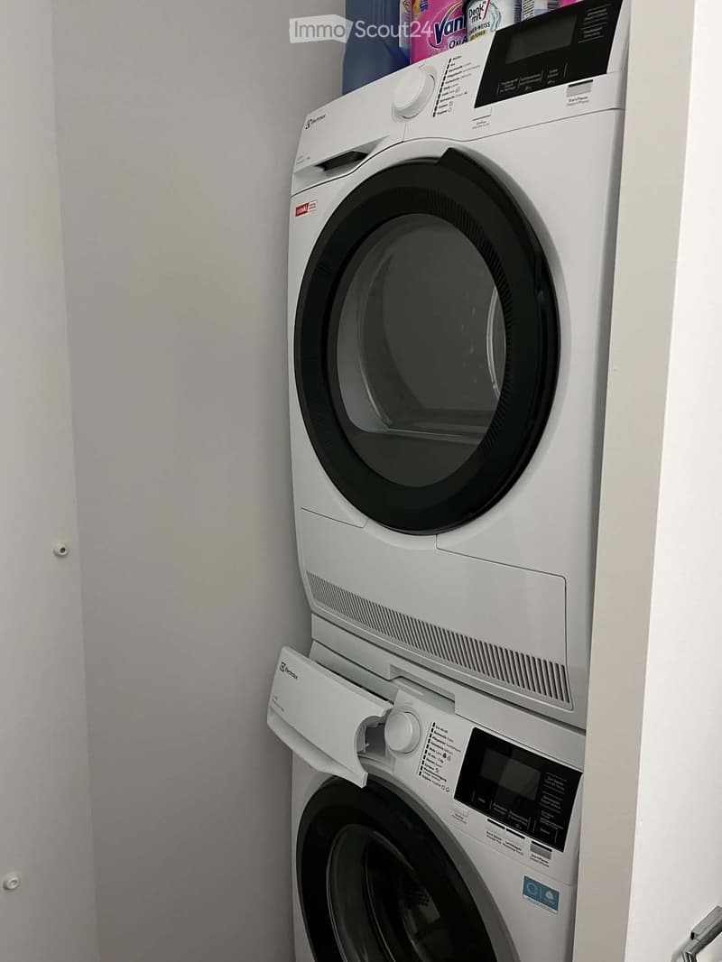 A compact, stacked washing machine and dryer unit in a bathroom corner, with detergent and cleaning supplies visible on top.