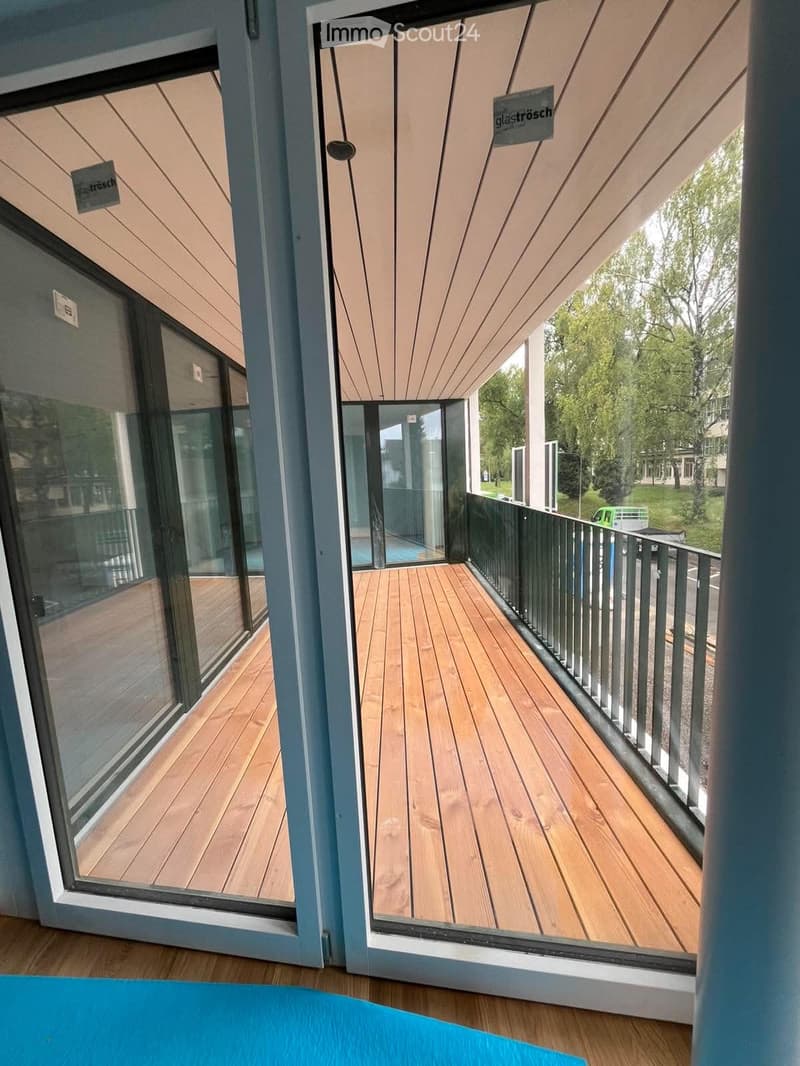 Modern wooden balcony with glass doors, metal railing, and a view of greenery and a street.