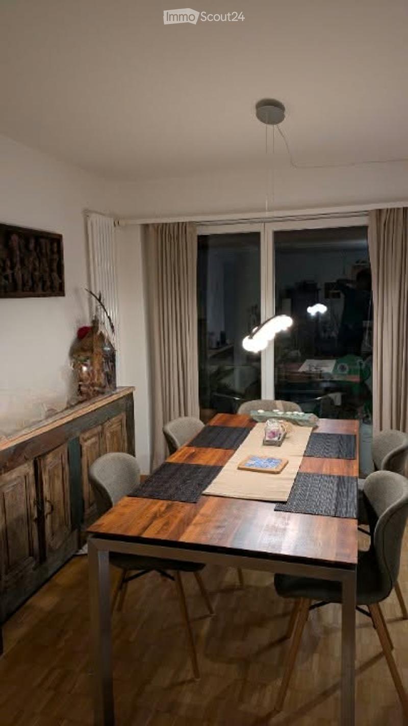 Dining room with a wooden dining table, six chairs, a cabinet with a picture frame, and a vase on top. Large windows with curtains provide natural light.