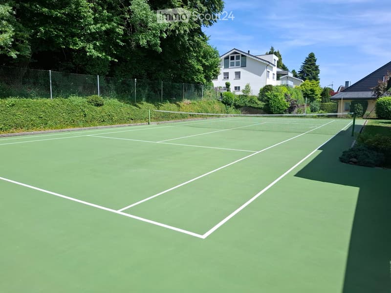 Tennis court with green surface and white lines, surrounded by a fence and trees, with houses visible in the background.