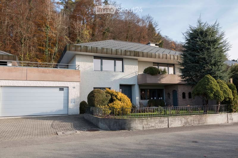 Modern two-story house with white exterior, grey roof, garage, and balconies surrounded by trees and shrubs.