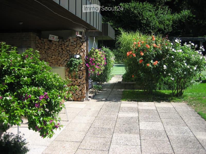Garden path lined with colorful flowers and shrubs, with a woodpile and a building in the background.