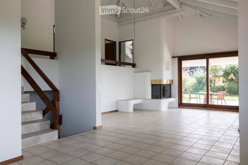 Bright, open hallway with tiled flooring, a staircase on the left, a fireplace in the center, and large glass doors leading to an outdoor area with chairs and a table.