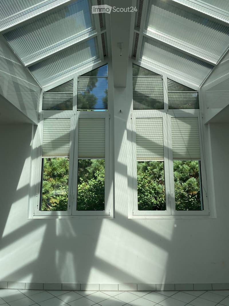 Bright, minimalist room with large windows and blinds, white tiled floor, and white walls. Natural light streams in, creating shadows on the walls.