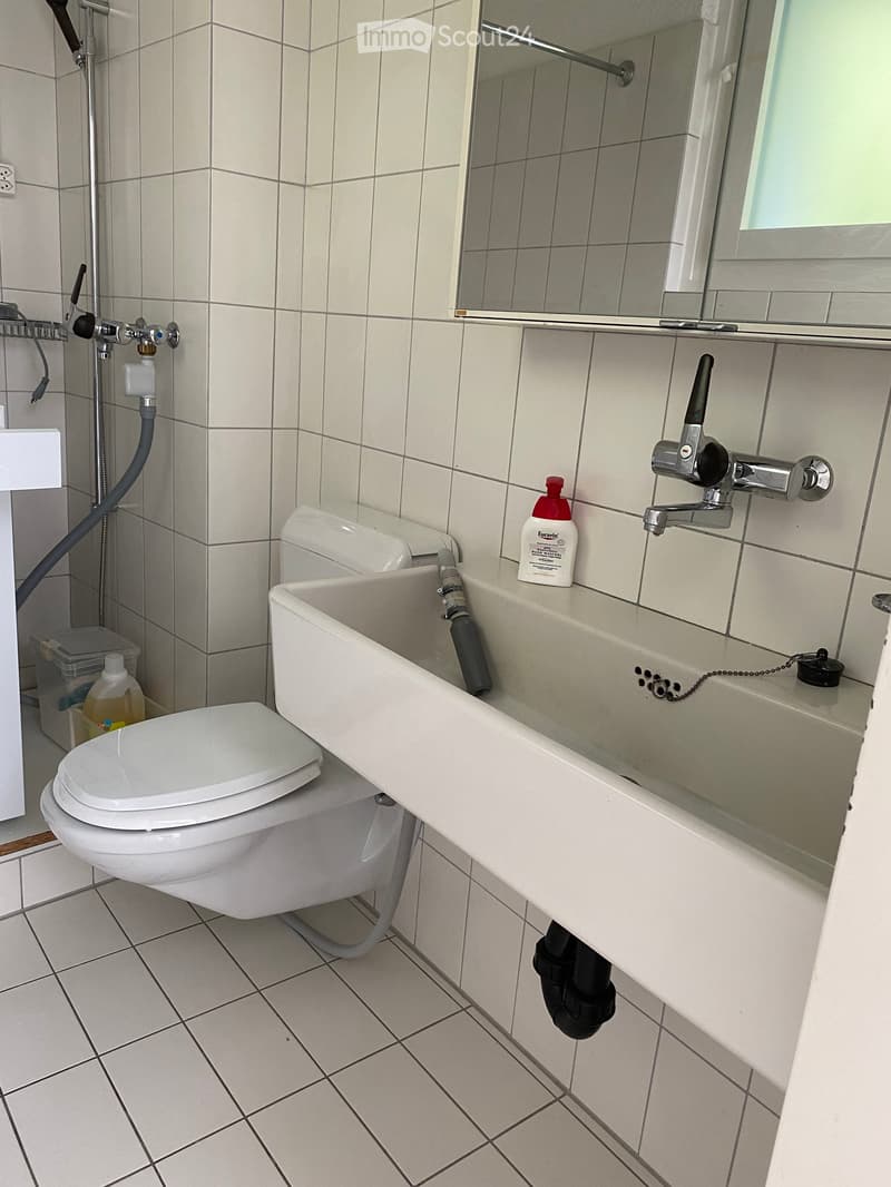 Bathroom with white tiled walls and floor, toilet, sink, mirror, and a bottle on the sink counter.