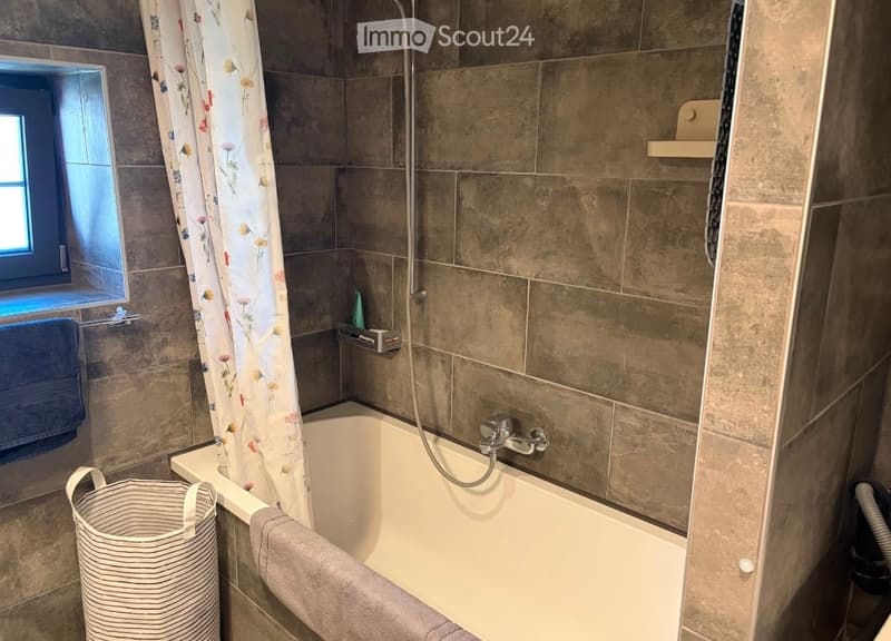 Modern bathroom with a bathtub, shower curtain, towels, a basket, and a window providing natural light.