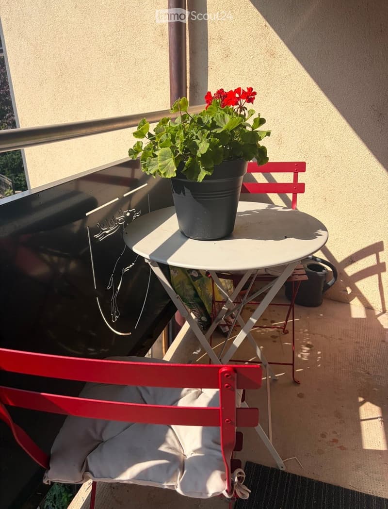 Balcony with a small round table, a red chair with a cushion, and a potted plant with red flowers.
