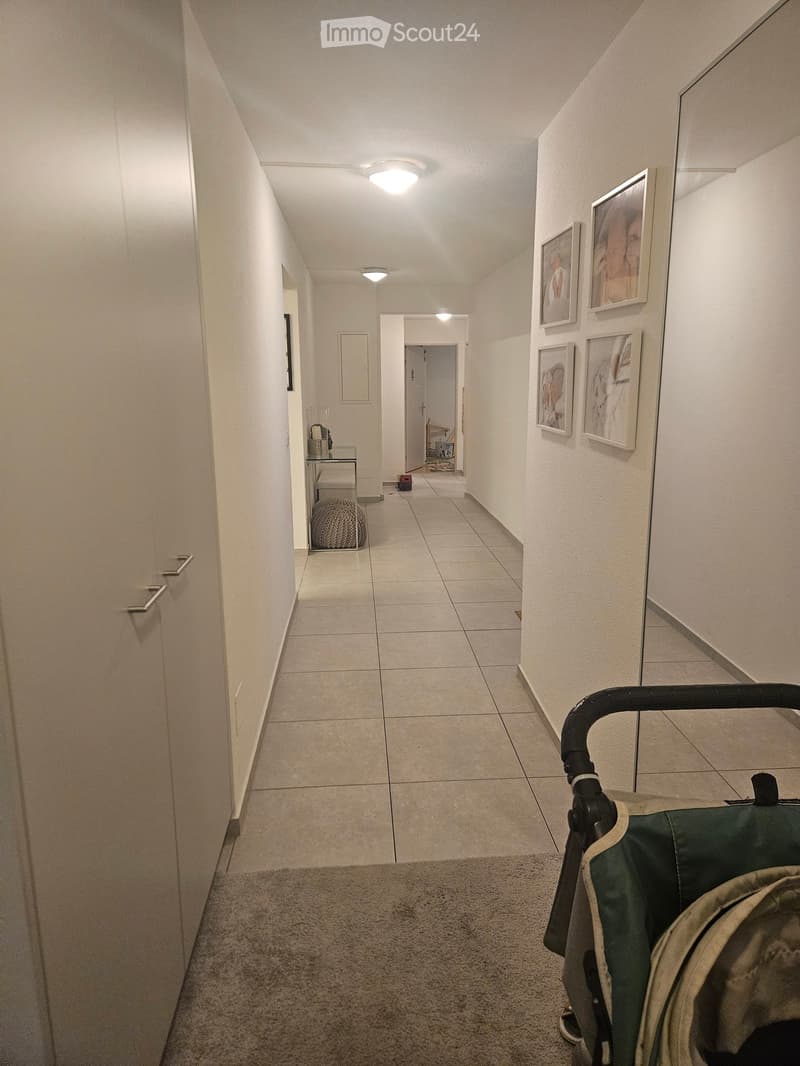A long, narrow hallway with tiled flooring, white walls, and several framed pictures on the right side. The hallway features recessed lighting and a small table with a chair in the background.