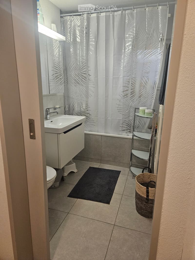 Modern bathroom with a white sink, toilet, bathtub, and shower curtain with leaf pattern. Includes a black bath mat, a small shelf with toiletries, and a woven basket on the floor.