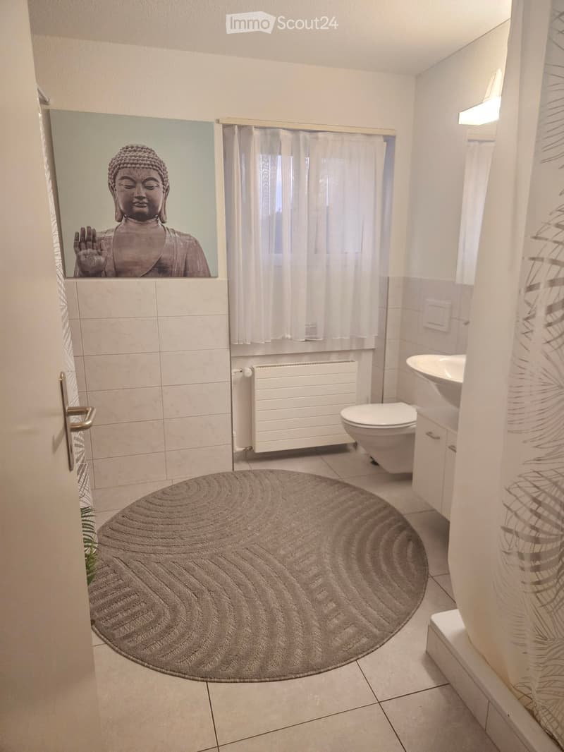Modern bathroom with white tiles, a round beige rug, a toilet, a sink, a mirror with a Buddha statue reflection, and a window with white curtains.