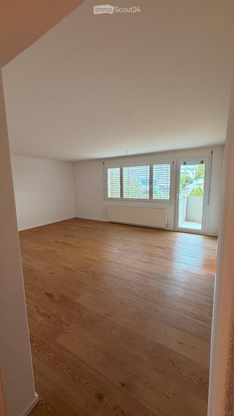 Empty room with wooden flooring, white walls, and large windows with blinds. A radiator is visible under the windows, and a sliding glass door leads to a balcony.