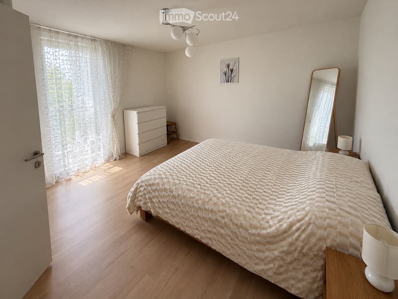 Cozy bedroom with wooden flooring, a neatly made bed, a nightstand with a lamp, a dresser, and a mirror. Light-colored curtains cover the window, allowing natural light to filter in.