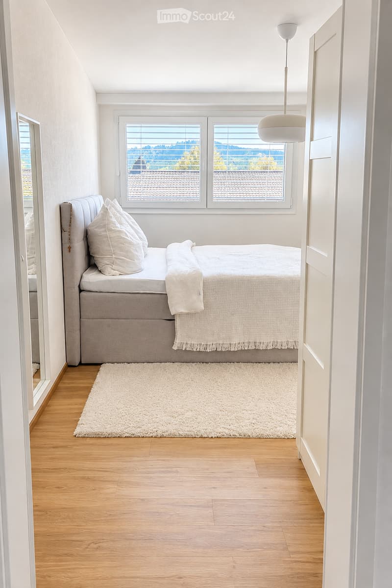 Modern bedroom with a large window offering a view, a neatly made bed with pillows and a blanket, a white shag rug, and a pendant light hanging from the ceiling.