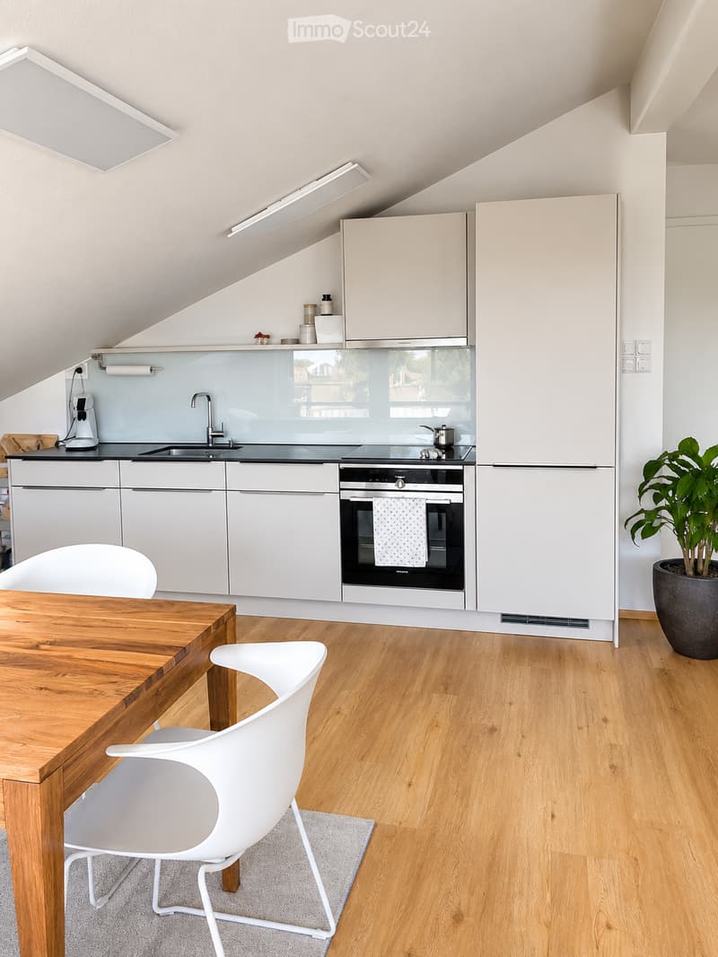 Modern kitchen with white cabinets, wooden flooring, a wooden dining table with white chairs, a sink, oven, refrigerator, and a potted plant in the corner.