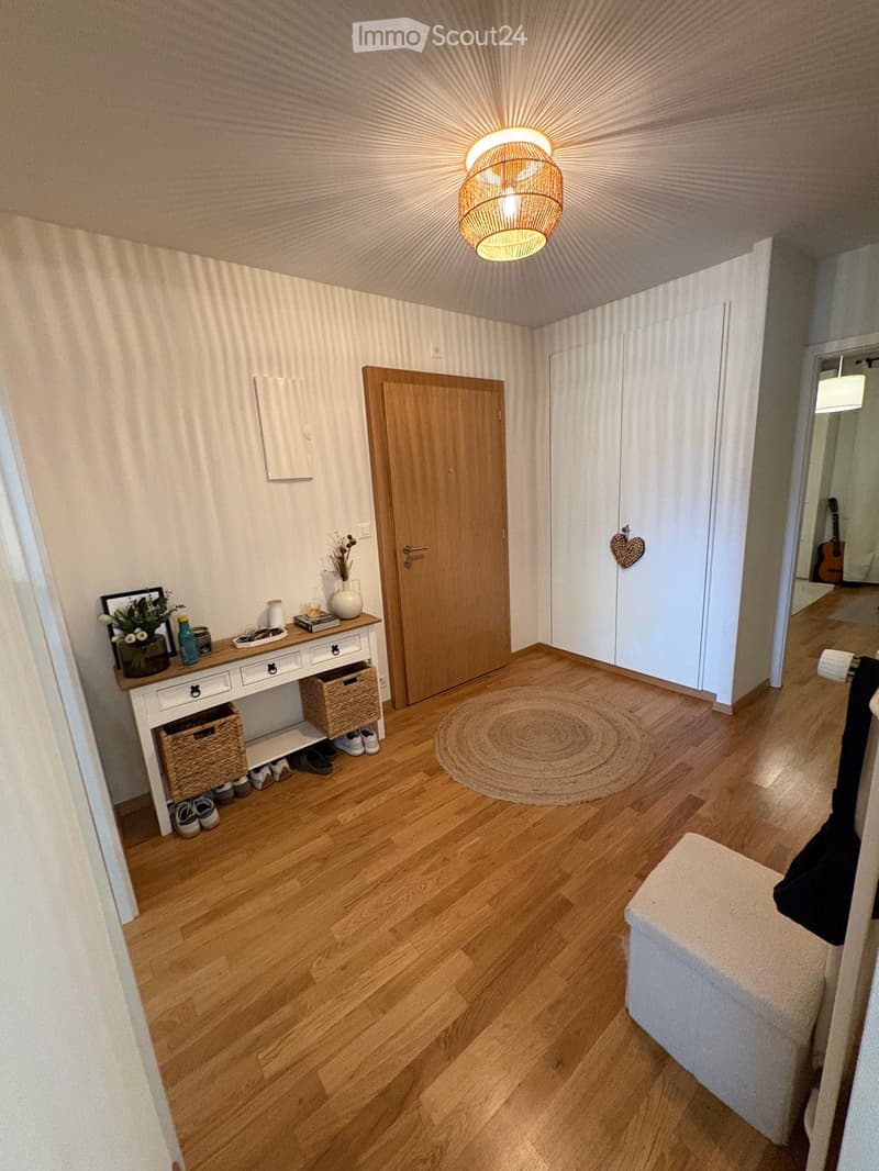 A well-lit hallway with wooden flooring, a round woven rug, a small console table with a vase and shoes, and a wooden door leading to another room.