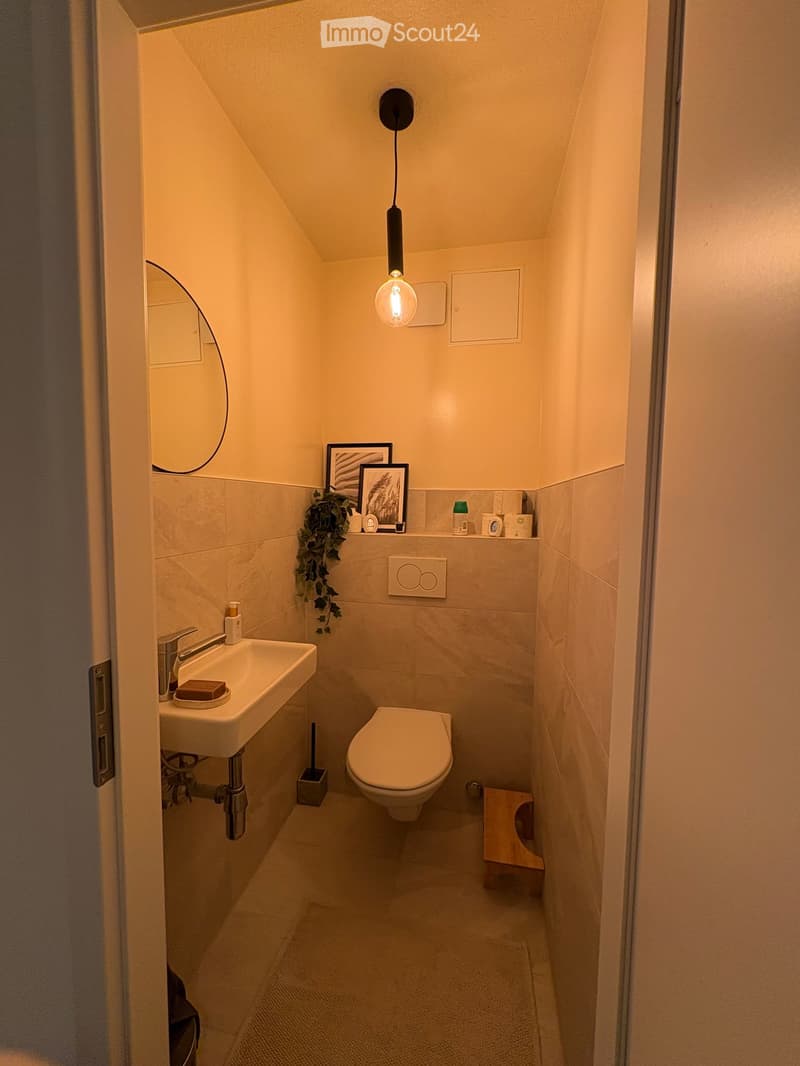 Compact bathroom with a white sink, toilet, and a small wooden stool. Features a modern mirror, framed pictures, and a hanging light fixture.