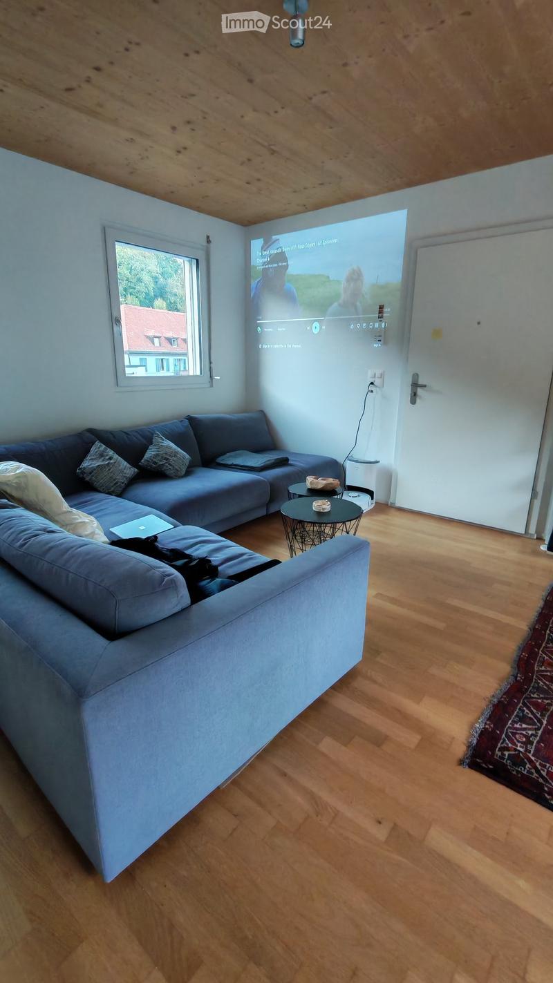 Modern living room with a large sectional sofa, coffee tables, a laptop, and a large screen displaying content. The room features wooden flooring and a cozy, contemporary design.