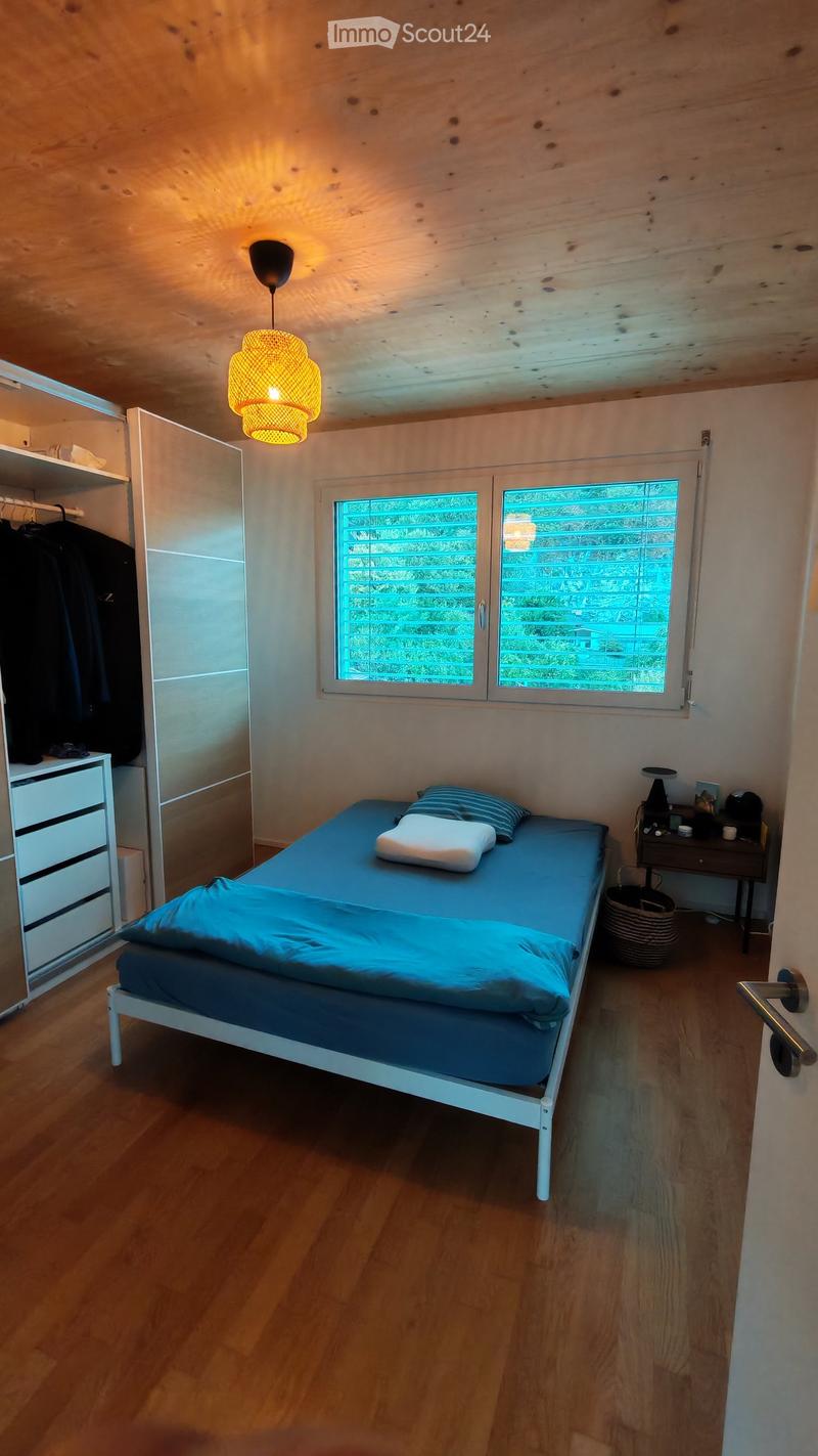 Cozy bedroom with a single bed, blue bedding, a nightstand with a lamp, a wardrobe, and a hanging woven basket. The room features wooden flooring and exposed wooden beams on the ceiling.
