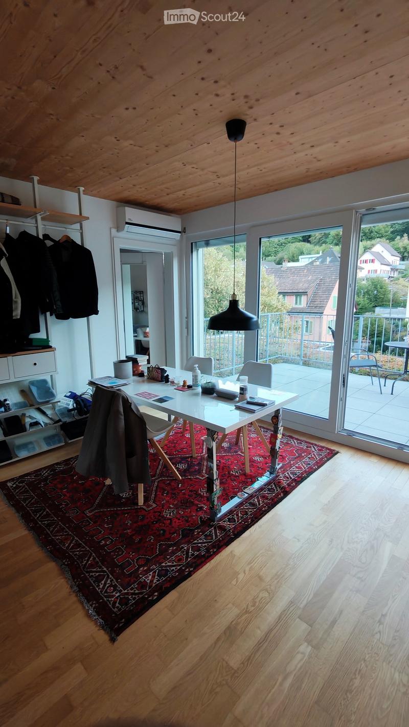 Bright room with wooden floors, a large red patterned rug, a white table with chairs, a hanging lamp, and a coat hanging on the wall. Large windows open to a balcony.