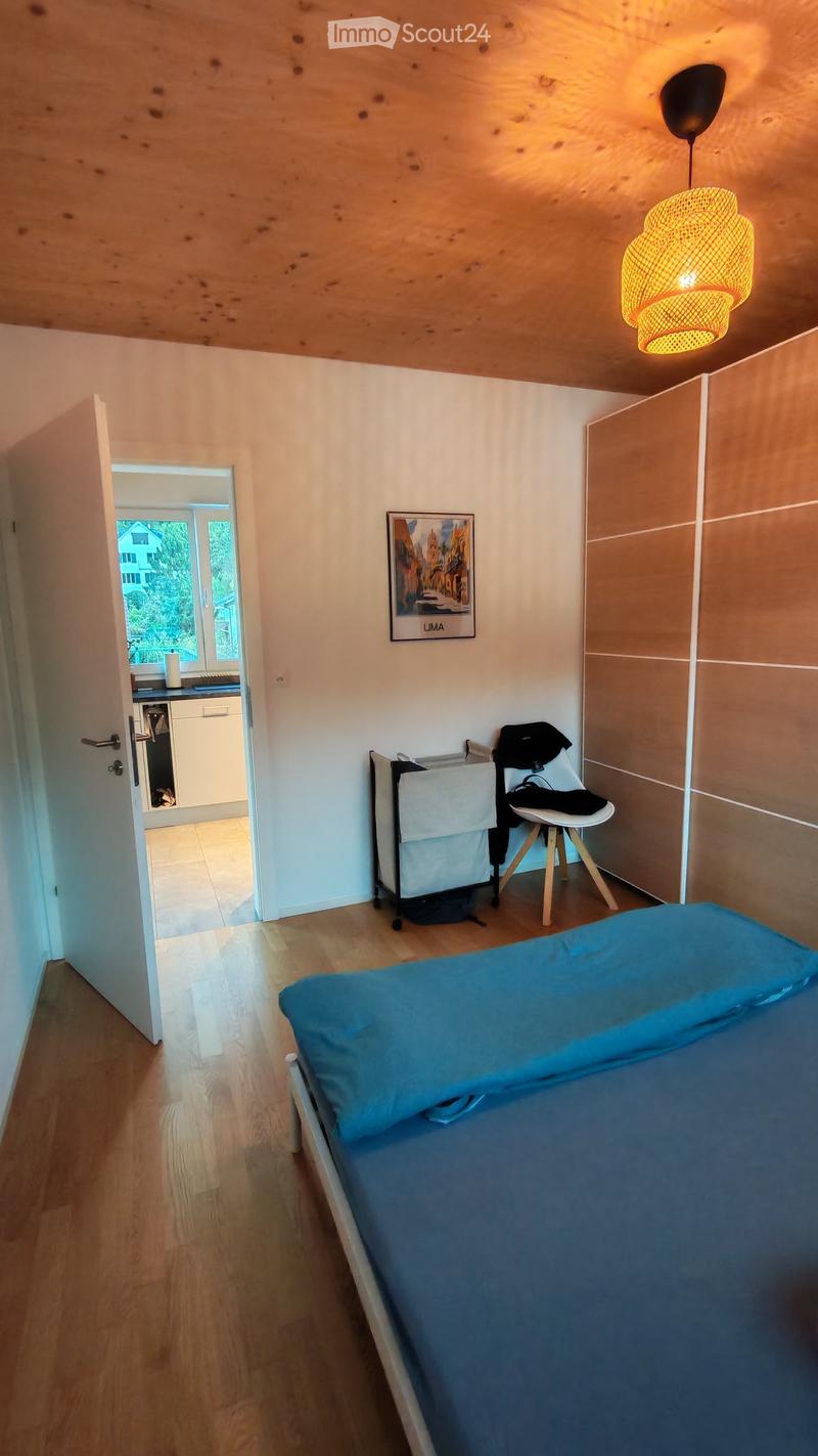 Cozy bedroom with wooden floor, bed with blue bedding, a chair, a picture frame on the wall, and a hanging lamp.