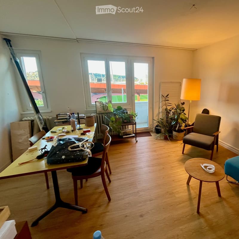 A multi-purpose room with a wooden floor, large windows, a table with chairs, a small round stool, a sofa, a floor lamp, and several potted plants.