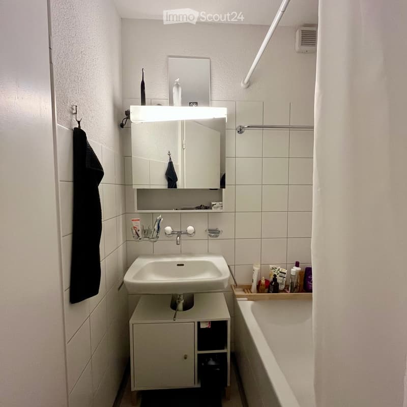 Modern bathroom with a white sink, mirror, cabinet, and a bath. The sink has a faucet and a small cabinet underneath. A towel hangs on the wall, and various toiletries are placed on the sink counter and a tray next to the bath.