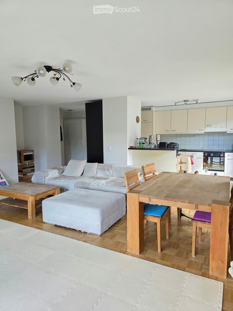 Bright, open-plan living and dining area with wooden flooring, white walls, and modern furniture including a sofa, ottoman, coffee table, dining table with chairs, and a small kitchen area with cabinets and appliances.