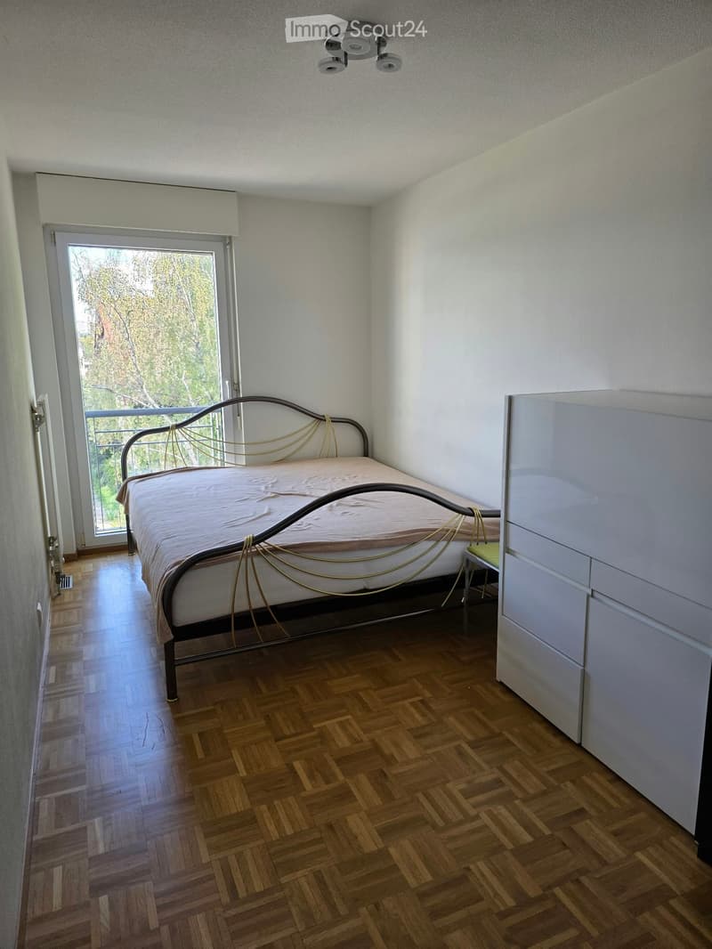 Bedroom with wooden herringbone flooring, a metal-framed bed with a beige bedspread, and a white wardrobe. A large window with blinds offers a view of greenery outside.