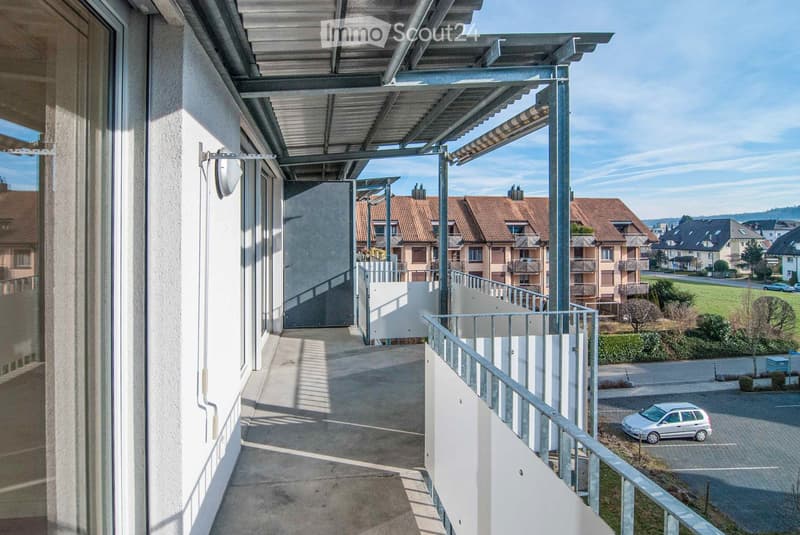 Balcony with a metal railing, covered walkway, and view of a residential area with houses, a parking lot, and a small car.