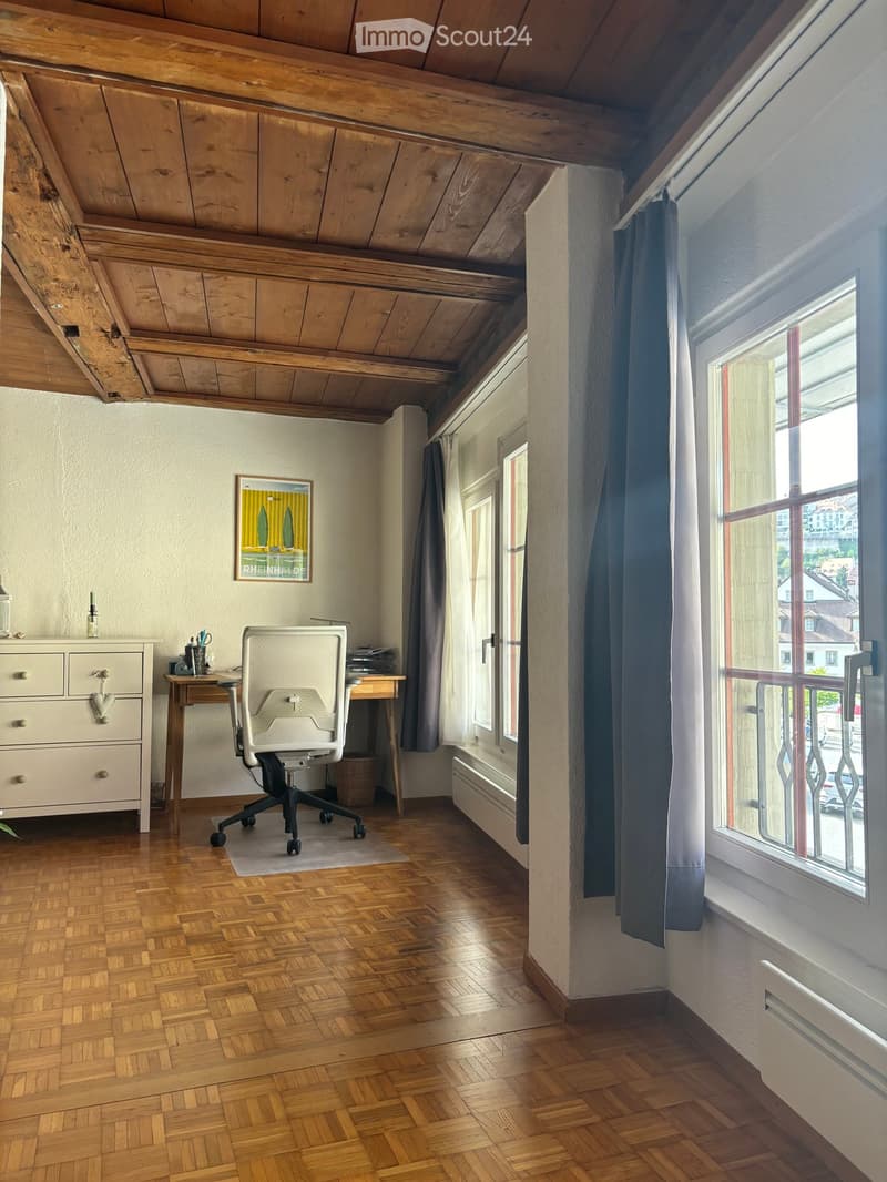 Bright room with wooden herringbone flooring, slanted wooden ceiling, large windows with dark curtains, a white office chair, a wooden desk, a small cabinet, and a framed picture on the wall.