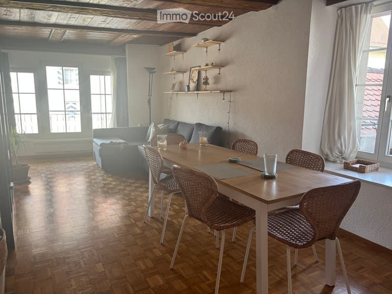 Bright living room with wooden flooring, large windows, a grey sofa, a dining table with four wicker chairs, and decorative shelves.