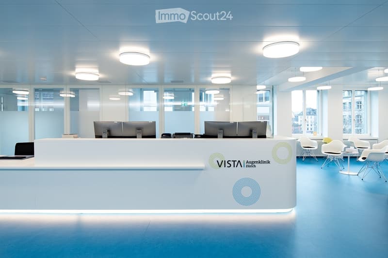 Modern office space with a reception desk, multiple computer screens, chairs, and tables arranged around the area.