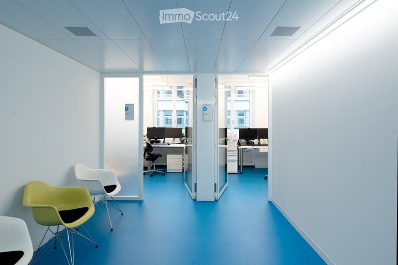 Modern office corridor with light blue walls and floor, featuring white and green chairs, desks with computers, and glass partitions.