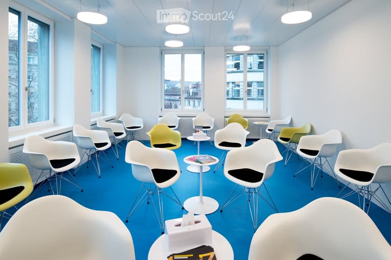 Bright, modern room with blue carpeting, white walls, and large windows. Multiple white and yellow chairs are arranged around small white tables. A tissue box and some books are placed on one of the tables.