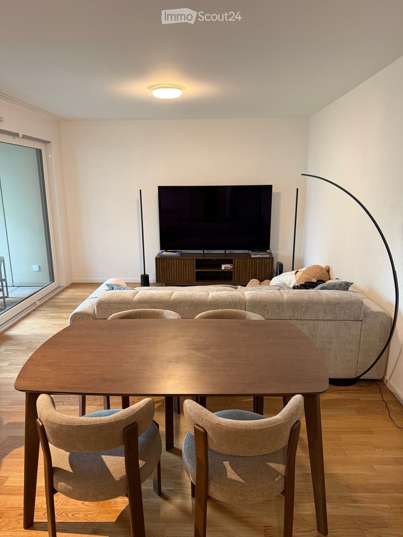 A modern living room with a grey sofa, a wooden dining table with two chairs, a television on a wooden stand, and a floor lamp near the sofa.