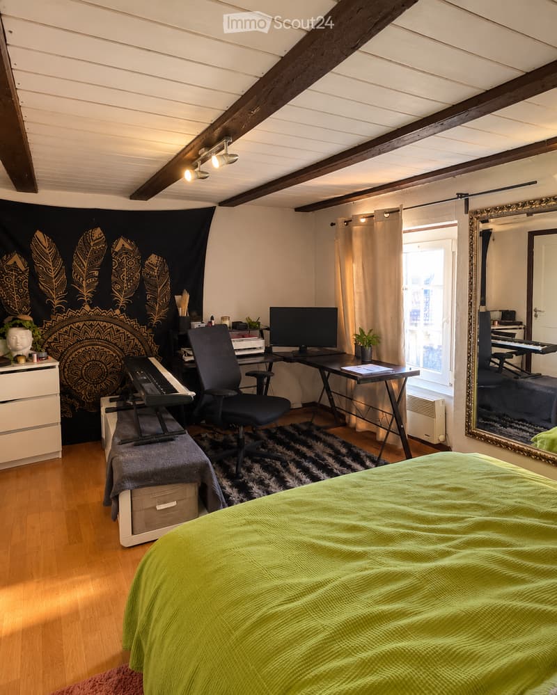 Cozy bedroom with a green bedspread, wooden floor, large mirror, desk with computer, chair, piano, and decorative elements including a tapestry and plants.