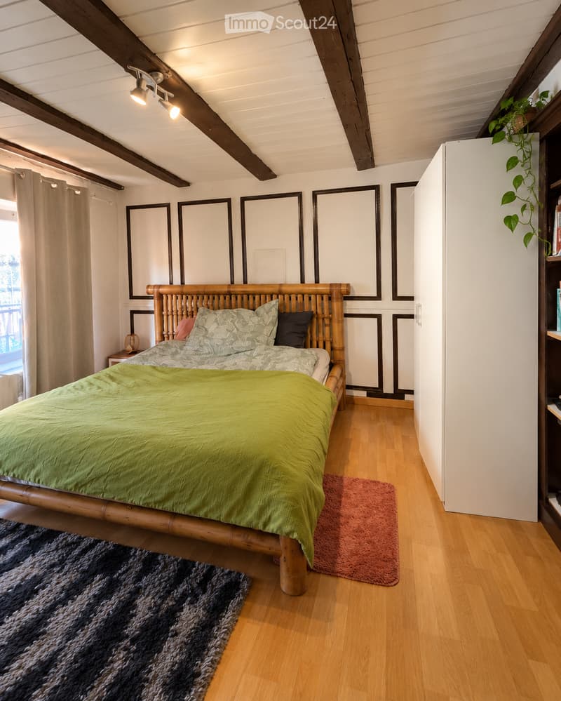 Cozy bedroom with wooden bed frame, green duvet, nightstand, mirrors, and rugs on wooden floor.