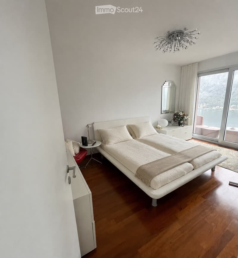 Modern bedroom with a large white platform bed, nightstand, chair, and a view of a lake through large windows.