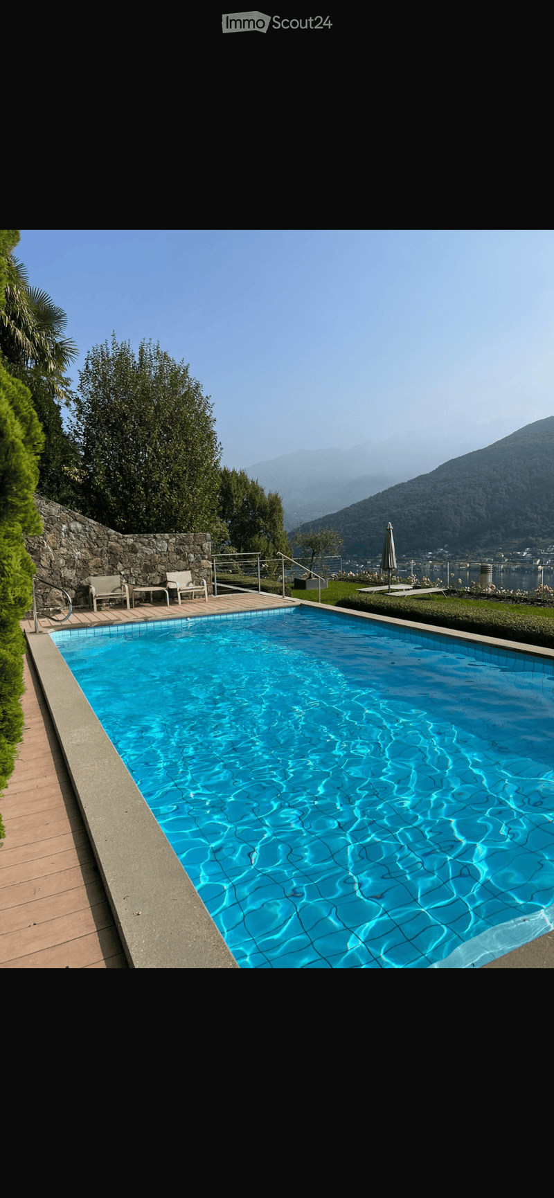 A rectangular swimming pool with clear blue water, surrounded by a stone wall, wooden decking, and outdoor furniture including chairs and a table. The background features a scenic mountain view and greenery.