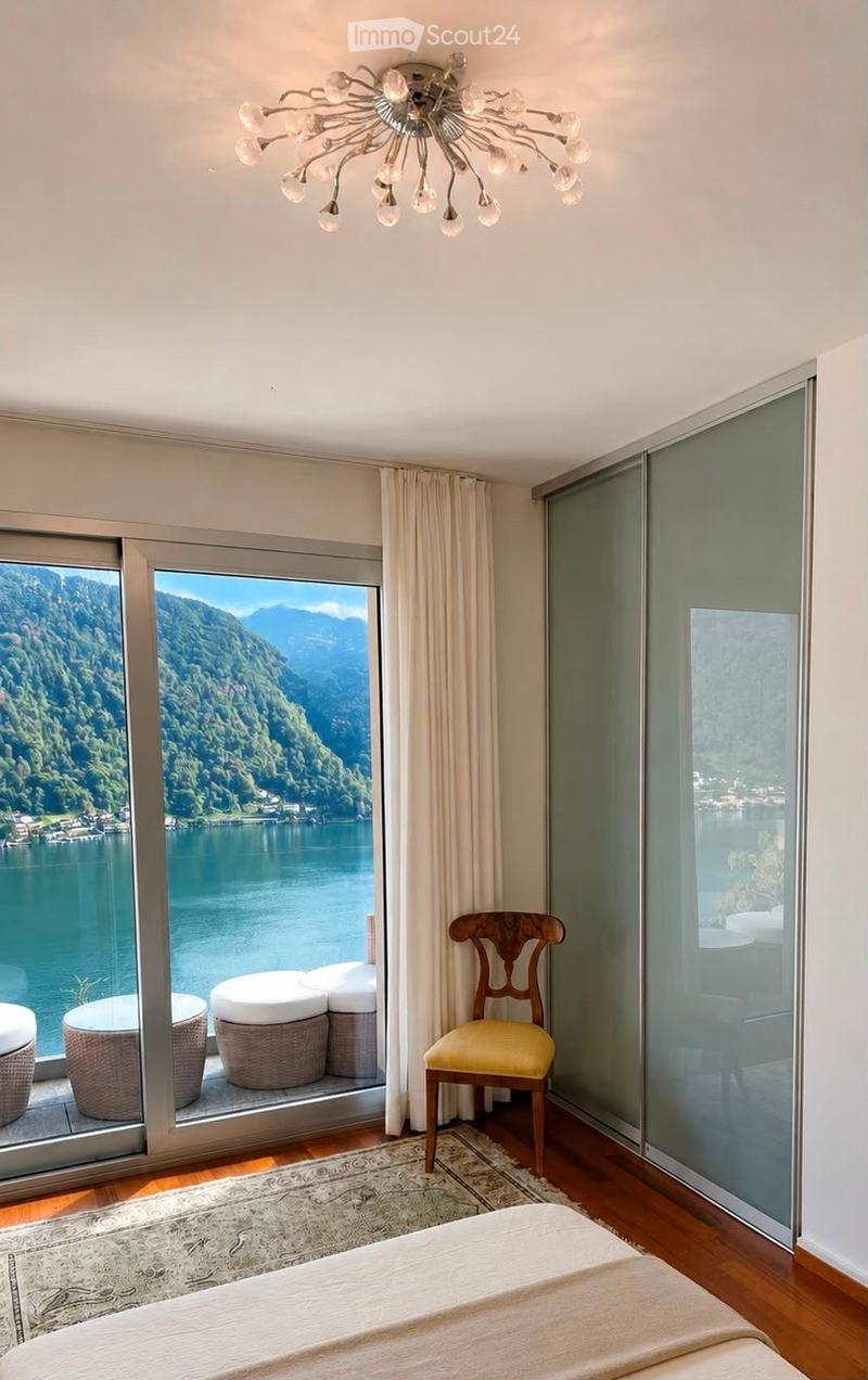 Elegant bedroom with a large window offering a scenic lake view, featuring a bed, a wooden chair with a yellow cushion, a chandelier, and a small seating area with stools on the balcony.