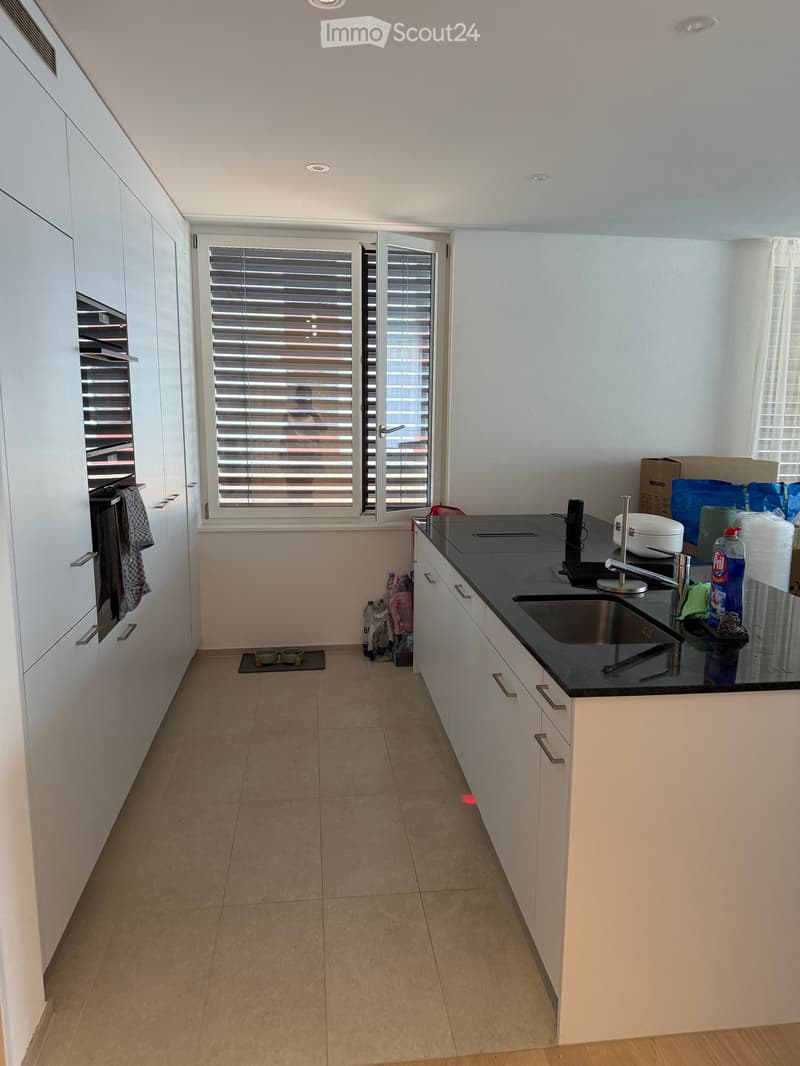 Modern kitchen with white cabinets, black countertop, stainless steel sink, and built-in oven. Features tiled floor and large window with blinds.