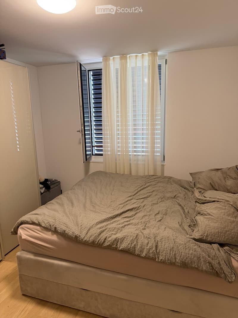 Bedroom with a large bed covered in beige bedding, a window with white curtains, and a wooden floor.