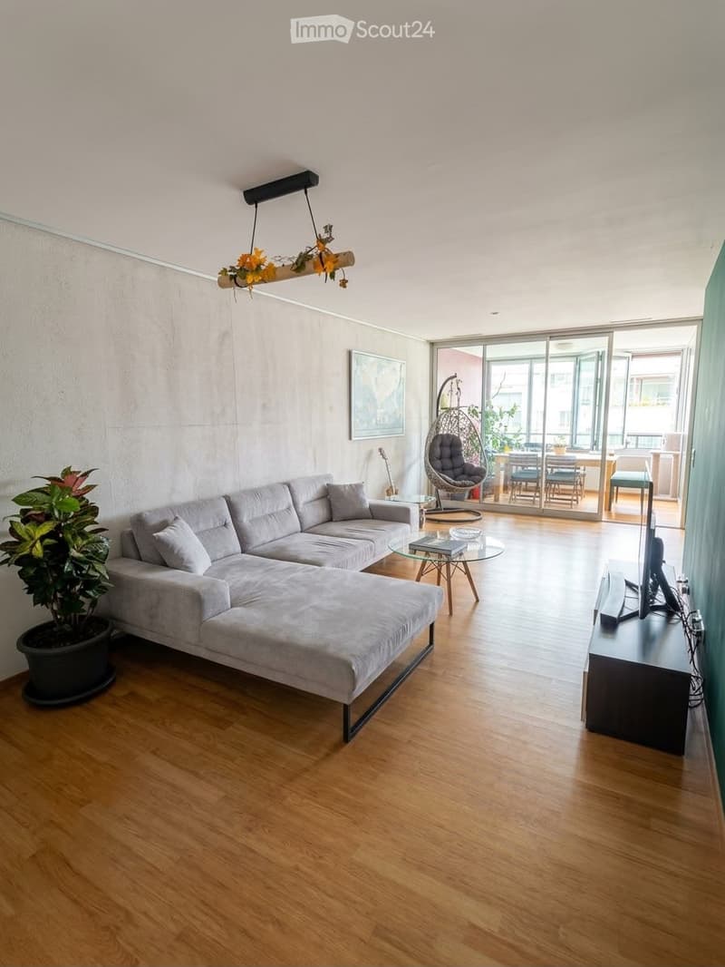 Modern living room with grey sectional sofa, coffee table, TV stand, wicker hanging chair, dining area with table and chairs, and a guitar leaning against the wall.