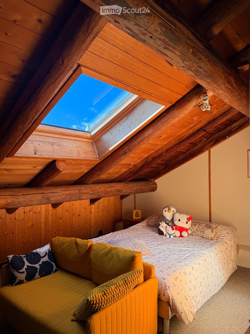 Cozy attic bedroom with wooden walls and exposed beams, featuring a large skylight, a yellow sofa, a bed with stuffed animals, and a nightstand with a picture frame.