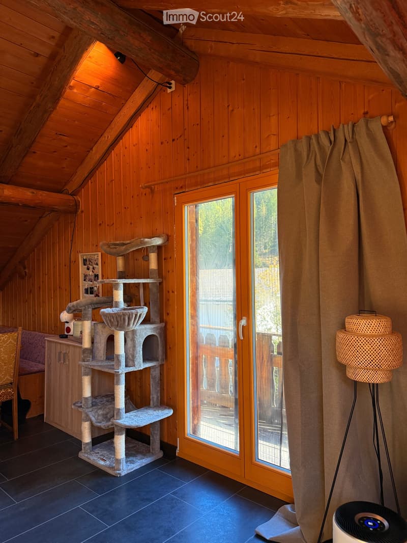 A cozy attic room with wooden walls and ceiling, featuring a multi-level cat tree, a large window with a view, a wicker lamp, and a fan on the floor.
