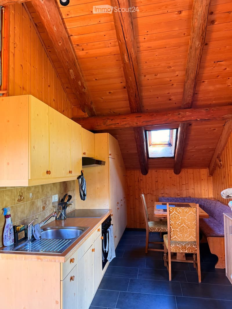 A cozy attic kitchen with wooden walls and ceiling, featuring a sink, oven, cabinets, a small dining area with a table and chairs, and a built-in sofa.