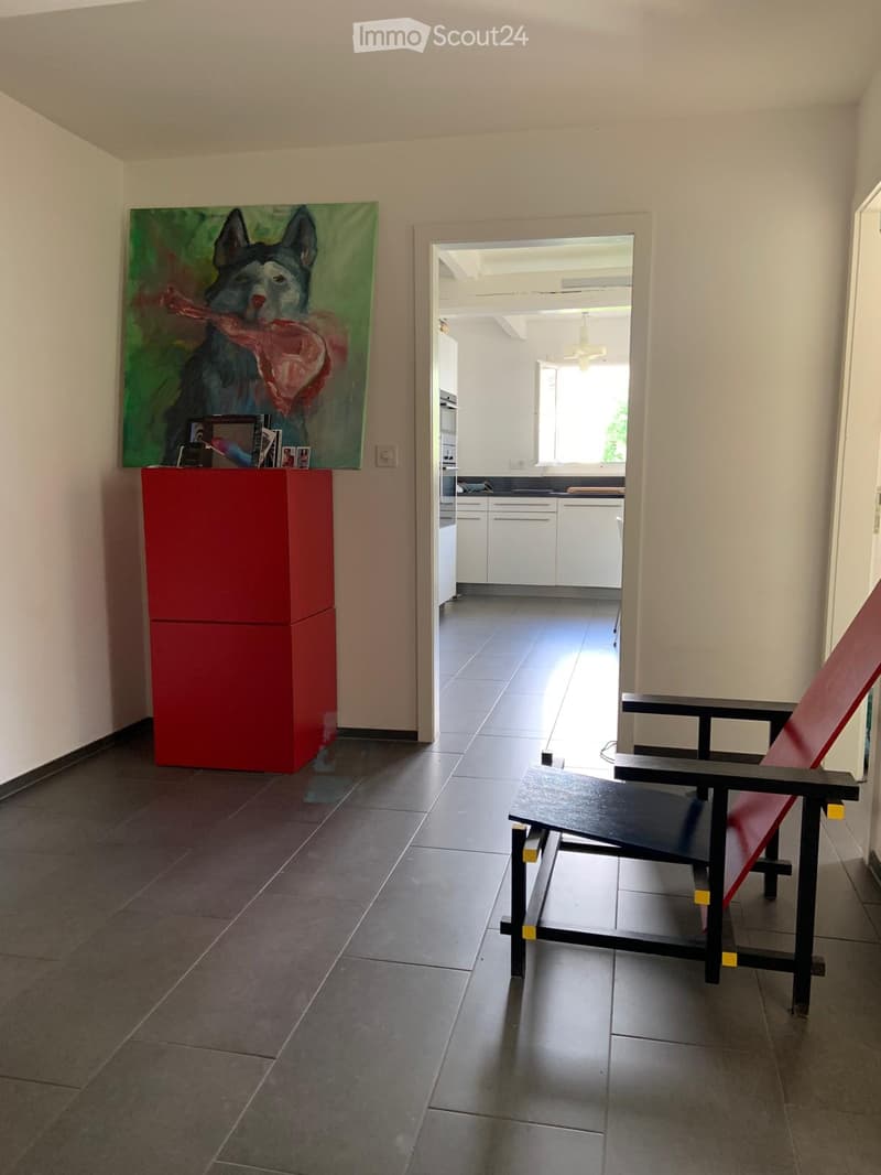 A modern, minimalist hallway with a red chair, a small table, a colorful painting, and a view into a bright kitchen area.