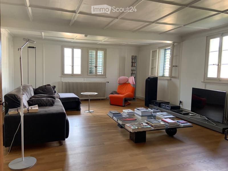 Living room with wooden flooring, large windows, a grey sofa, an orange armchair, a coffee table with books, a TV, and a floor lamp.